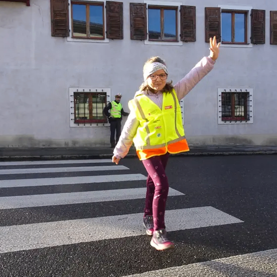 Ein junges Mädchen in einer reflektierenden Weste winkt, während sie die Straße an einem Zebrastreifen überquert. Hinter ihr steht ein Verkehrspolizist.