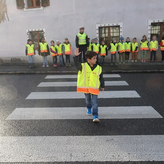 Ein Junge in einer reflektierenden Weste steht auf einem Zebrastreifen, winkt und hat eine Reihe von Kindern und einen Erwachsenen in einer Polizeimütze hinter sich.