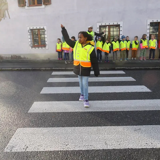 Ein Kind in einer reflektierenden Weste steht auf einem Zebrastreifen und zeigt mit einer Hand, während mehrere Kinder in Westen zusehen.