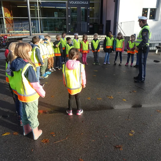 Eine Gruppe von Kindern in Reflektorwesten steht vor einem Gebäude, wobei ein Polizeibeamter mit ihnen spricht.