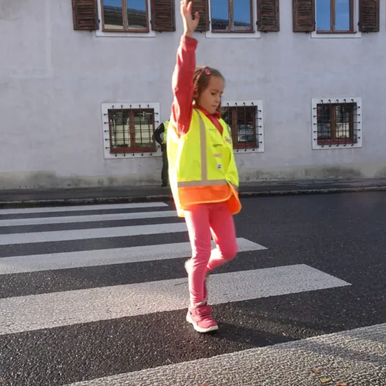 Ein junges Mädchen in einer reflektierenden Weste überquert die Straße und winkt mit der Hand. Sie befindet sich auf einem Zebrastreifen mit weißen Streifen. Hinter ihr steht ein Gebäude mit mehreren Fenstern.