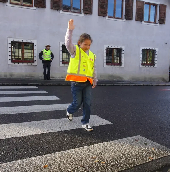 Ein junges Mädchen in einem gelben Weste winkt vor der Kamera auf der Straße. Ein Verkehrswächter steht hinter ihr, ebenfalls in einer gelben Weste. Die Szene spielt sich vor einem Gebäude mit mehreren Fenstern ab.