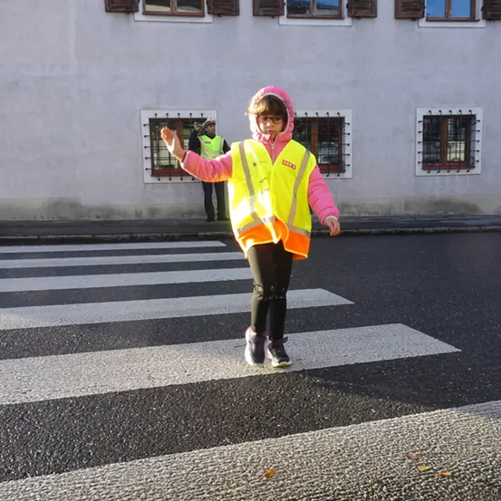 Ein Kind in einer Warnweste überquert einen Zebrastreifen vor einem Gebäude mit mehreren Fenstern. Hinter ihm steht eine Person in einer Warnweste in der Nähe des Gebäudes.
