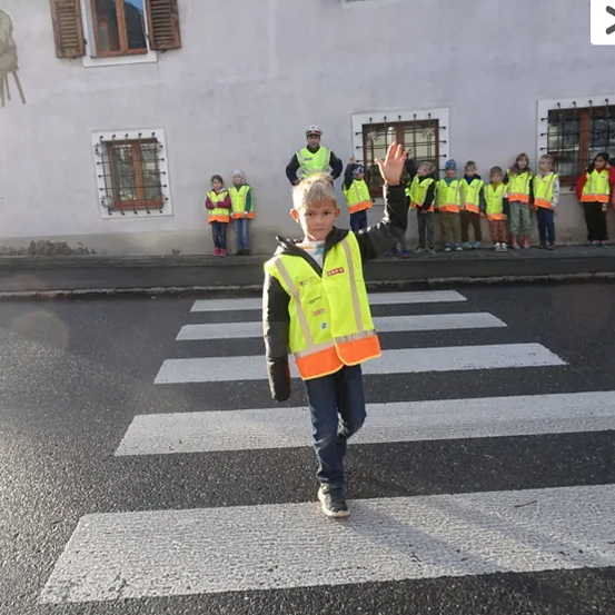 Ein junger Junge in einer reflektierenden Weste geht über einen Zebrastreifen. Hinter ihm steht eine Reihe von Kindern in gelben Westen auf dem Bürgersteig, und ein Erwachsener in einer gelben Weste winkt. Ein Gebäude mit mehreren Fenstern ist im Hintergrund.