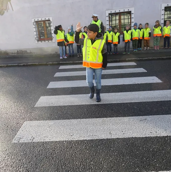 Ein junger Mensch in einer Sicherheitsweste leitet den Verkehr auf einer Straße, wobei Kinder und Erwachsene in reflektierenden Westen hinter ihnen stehen.