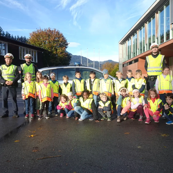 Eine Gruppe von Kindern und zwei Polizeibeamten in Warnwesten posiert für ein Foto vor einem Auto. Im Hintergrund befinden sich Gebäude und Bäume.