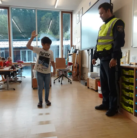 Ein Polizeibeamter demonstriert einem Kind in einem Klassenzimmer mit großen Fenstern und einer Gitarre in der Ecke eine Sicherheitsprozedur.