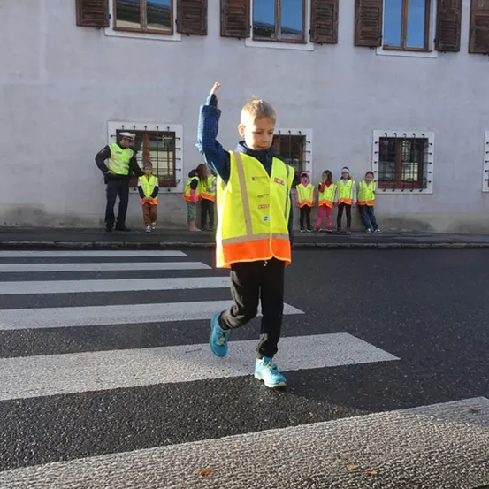 Ein Kind in einer gelben Sicherheitsweste überquert die Straße vor einem Gebäude mit mehreren Fenstern. Andere Kinder und Erwachsene in gelben Westen stehen auf dem Bürgersteig.