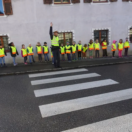 Ein Polizist steht auf einem Zebrastreifen, mit Kindern in reflektierenden Westen hinter ihm. Sie stehen vor einem Gebäude mit Fenstern.