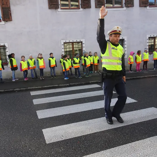 Ein Verkehrspolizist signalisiert Kindern in Warnwesten, die Straße zu überqueren. Sie stehen in einer Reihe auf dem Bürgersteig vor einem Gebäude.