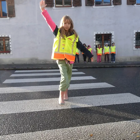 Ein kleines Mädchen mit Warnweste geht über einen Zebrastreifen. Hinter ihr stehen mehrere Kinder in Warnwesten in der Nähe eines Gebäudes mit mehreren Fenstern.