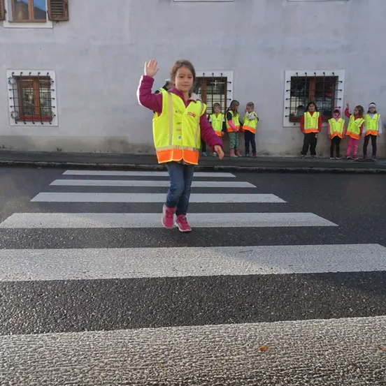 Ein junges Mädchen in einer gelben Weste winkt, während es eine Straße überquert. Dahinter stehen mehrere Kinder in gelben Westen auf dem Bürgersteig.