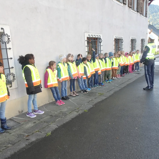 Kinder in gelben Westen stehen in einer Reihe vor einem Gebäude, ein Polizist steht am Ende.