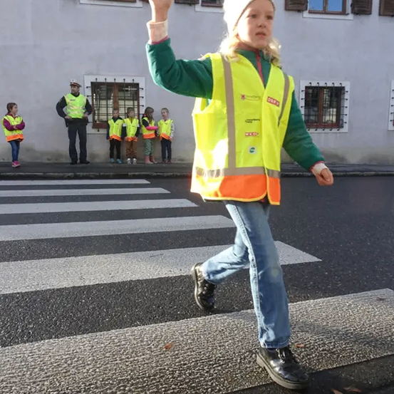 Ein Mädchen in einer Sicherheitsweste geht mit erhobener Hand über einen Zebrastreifen. Dahinter stehen ein Mann und mehrere Kinder in Sicherheitswesten auf dem Bürgersteig.