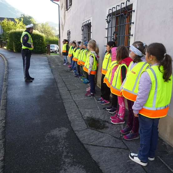 Ein Polizist in einer Warnweste instruiert eine Reihe von Kindern in passenden Westen, die auf einem Bürgersteig stehen.