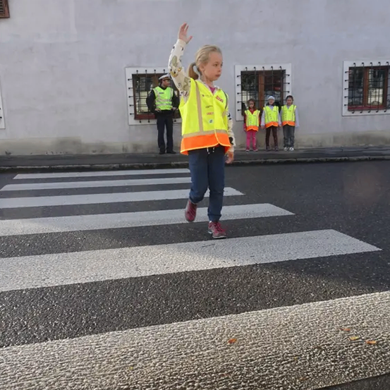 Ein junges Mädchen in einem gelben Warnweste winkt mit der Hand auf einem Zebrastreifen. Dahinter steht ein Gebäude mit Fenstern und einer Gruppe von Kindern in gelben Warnwesten.