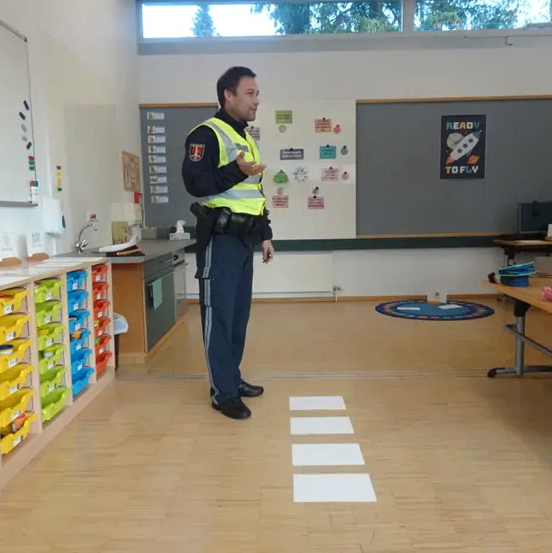 Ein Polizist steht in einem Klassenzimmer, trägt eine Warnweste und spricht. Eine Tafel und verschiedene Poster hängen an der Wand.