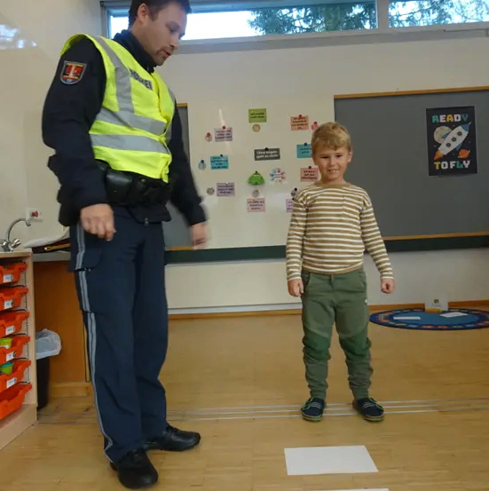 Ein Polizist steht in einem Klassenzimmer und trägt eine gelbe Weste. Ein junger Junge steht neben ihm und schaut den Polizisten an. Der Raum hat eine weiße Wand, eine Tafel und einen runden Teppich.