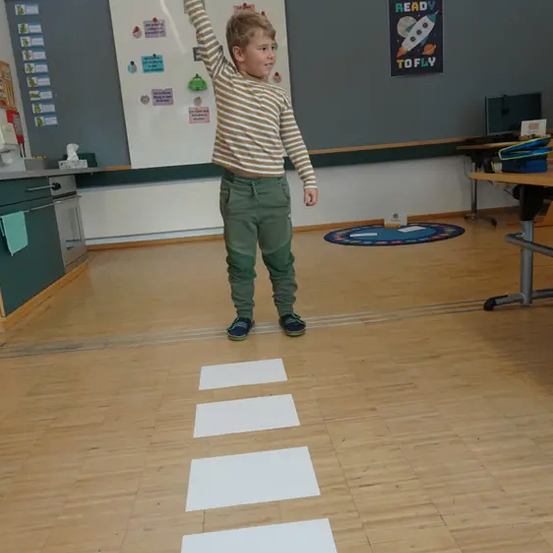 Ein junger Junge steht auf einem Holzboden, hebt seine rechte Hand und trägt ein gestreiftes Hemd und grüne Hose. Vier weiße Quadrate sind auf dem Boden.