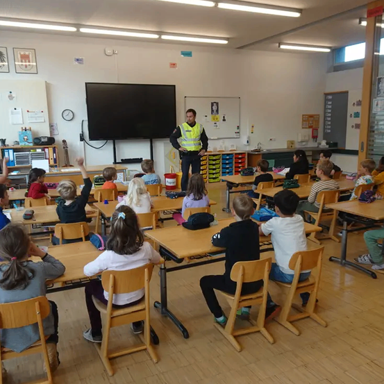 Ein Klassenzimmer mit Schülern und einem Polizeibeamten, eine Tafel und ein Monitor sind an der Vorderseite des Raumes.