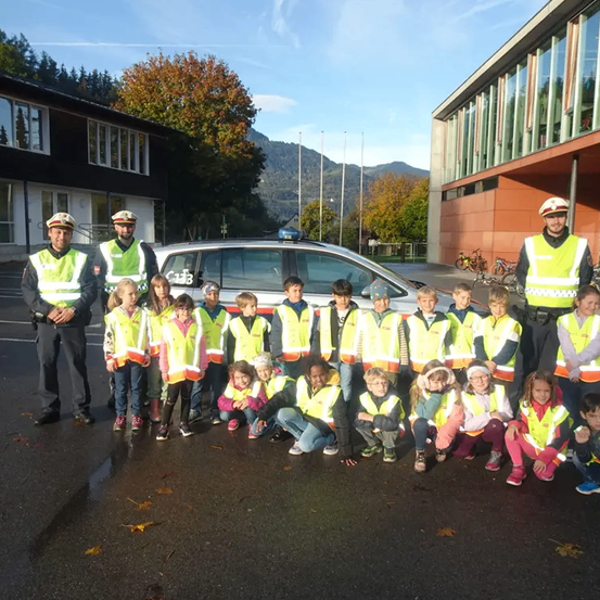 Eine Gruppe von Kindern in gelben Westen posiert für ein Foto mit zwei Polizisten und einem Polizeiauto im Hintergrund.