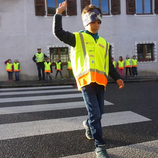 Ein Junge in einer reflektierenden Weste winkt, während er die Fußgängerfurt überquert. Dahinter stehen mehrere Kinder in reflektierenden Westen und ein Mann in Uniform auf dem Bürgersteig.
