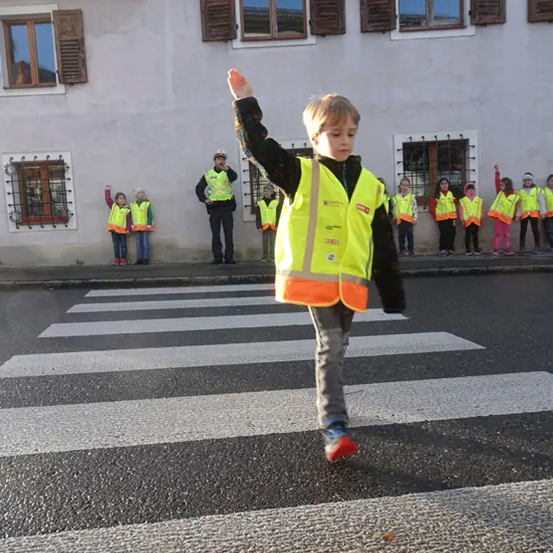 Ein junger Junge in einem neongelben Warnweste winkt mit der Hand, während er auf einem Zebrastreifen steht. Hinter ihm stehen andere Kinder in gelben Westen, ein Mann in einem Hut und ein Gebäude mit mehreren Fenstern.