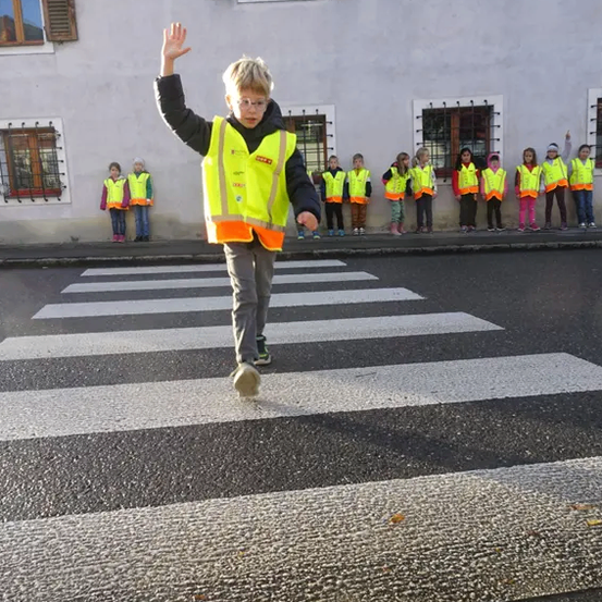 Ein Junge in einer gelben Warnweste geht auf einem Zebrastreifen und winkt zur Kamera. Dahinter steht eine Gruppe von Kindern in Warnwesten auf dem Bürgersteig.