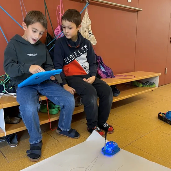 Zwei Jungen sitzen auf einer Bank, einer schaut auf ein Tablet, während der andere einem Roboter zusieht, wie er auf einem weißen Papier zeichnet.