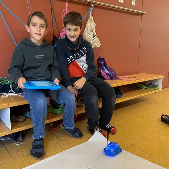 Zwei junge Jungen sitzen auf einer Bank, einer mit einem Tablet und der andere mit einer Zeichnung. Sie sind in einem Raum mit gefliestem Boden und einer Tafel.