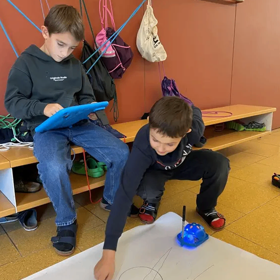 Zwei Jungen arbeiten auf einem Boden, einer zeichnet mit einem Stift auf einem weißen Papier und der andere sitzt auf einer Bank mit einem Tablet in den Händen.