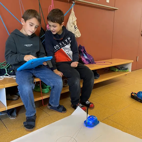 Zwei Jungen sitzen auf einer Bank, jeder hält ein Tablet. Ein Junge lächelt und schaut auf das Tablet, der andere konzentriert sich ebenfalls darauf. Sie befinden sich in einem Raum mit Holzboden und einer Wand mit hängenden Gegenständen.