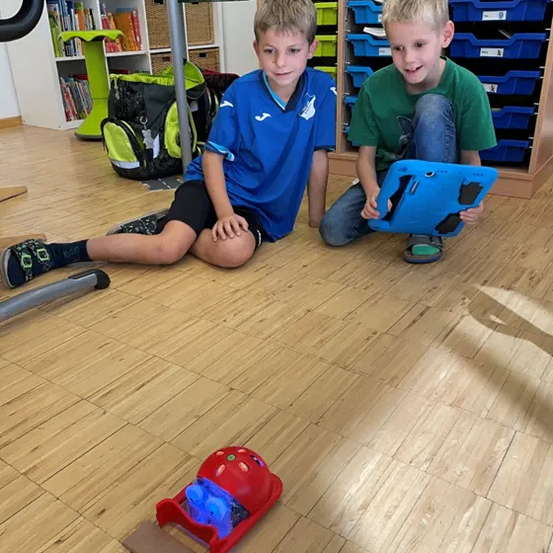 Zwei Jungen sitzen auf einem Holzboden und benutzen ein Tablet. Ein kleines rotes Spielzeug liegt vor ihnen auf dem Boden.