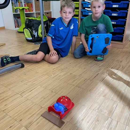Zwei Jungen sitzen auf dem Boden in einem Klassenzimmer, einer hält ein Tablet. Ein roter Spielzeugwagen steht vor ihnen auf dem Boden. Hinter ihnen steht ein Schreibtisch mit einem Regal mit Büchern und Aufbewahrungsboxen.