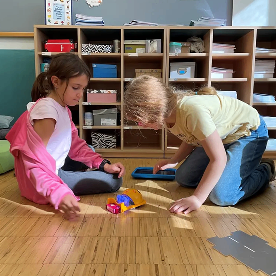 Zwei Mädchen sitzen auf dem Boden, eine spielt mit einem Tablet, die andere spielt mit einem Spielzeug auf einem Holzboden, in der Nähe eines Holzregals mit Büchern und Kisten.