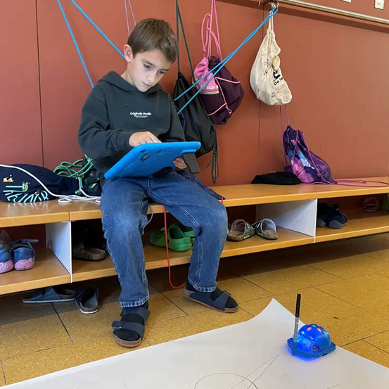 Ein junger Junge sitzt auf einer Bank, in ein Tablet vertieft, mit einem Roboter in der Nähe. Verschiedene Taschen und Schuhe hängen und sind um ihn herum platziert.