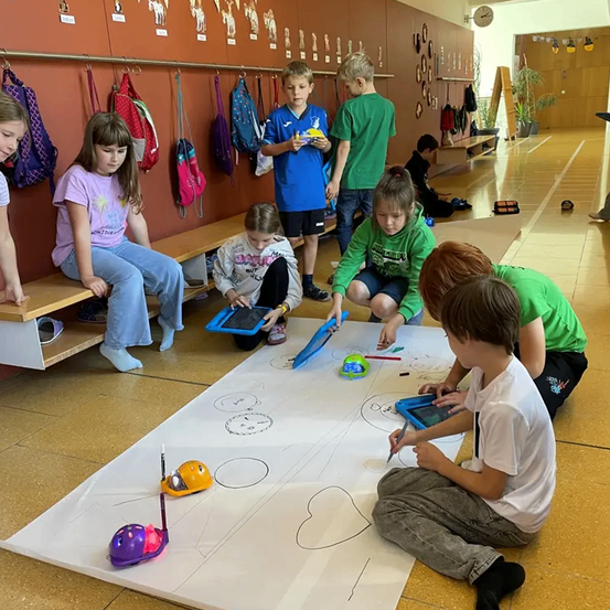 Kinder sitzen um ein großes Blatt Papier herum, zeichnen und benutzen Tablets. Sie sitzen auf dem Boden, während Rucksäcke an der Wand hinter ihnen hängen.