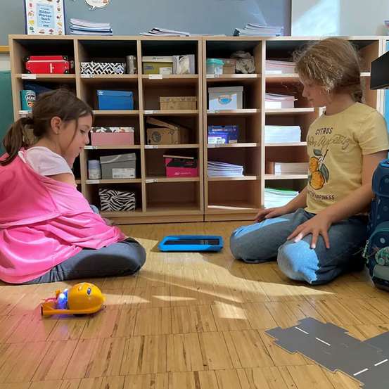 Zwei Mädchen sitzen auf dem Boden und sehen sich an. Eines trägt eine pinke Jacke und das andere ein gelbes T-Shirt. Sie spielen mit einem Spielzeugauto auf dem Boden. Dahinter steht ein hölzernes Regal mit verschiedenen Gegenständen.