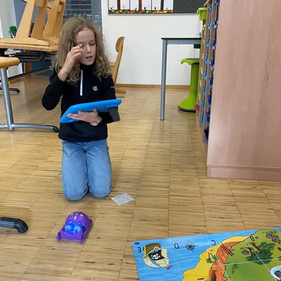 Ein junges Mädchen in einem Klassenzimmer, kniet und hält ein blaues Tablet, scheint auf etwas konzentriert zu sein. Eine bunte Karte liegt auf dem Boden. Stühle und ein Schreibtisch sind im Hintergrund.