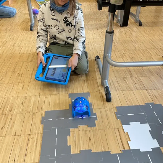 Ein junges Mädchen spielt mit einem Roboter auf einem Holzboden und trägt ein weißes Sweatshirt mit verschiedenen Aufdrucken.