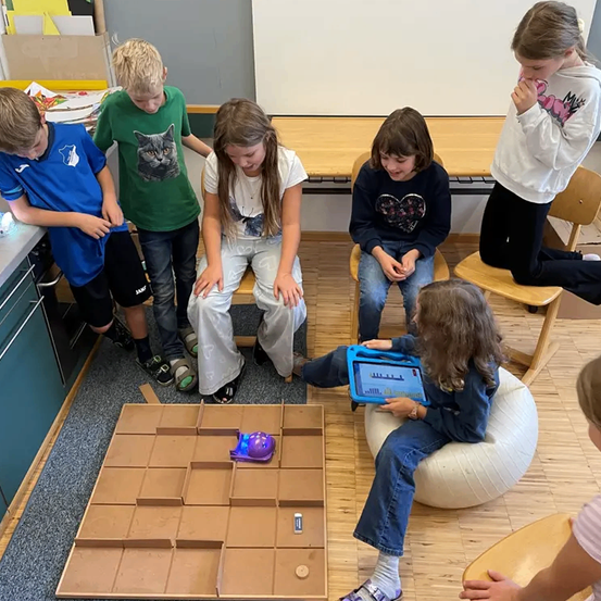 Eine Gruppe von Kindern spielt ein Spiel mit einem Roboter auf einem Holzbrett. Ein Kind steuert den Roboter mit einem Tablet.