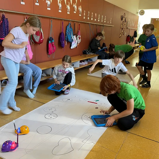Mehrere Kinder arbeiten in einem Klassenzimmer an einer Zeichnung. Ein Junge sitzt mit einem Tablet auf dem Boden. Ein Mädchen steht mit einem Tablet. Ein Junge steht und hält einen Ball.