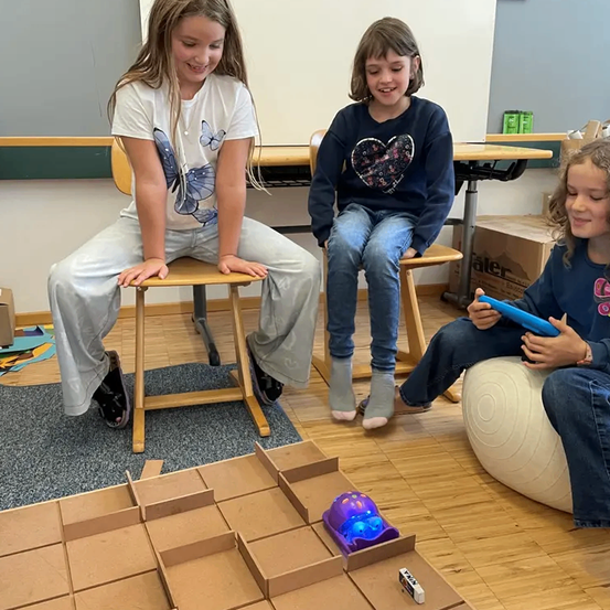 Drei Mädchen sitzen auf Stühlen um ein Labyrinth mit einem kleinen Roboter. Ein Mädchen hält ein Tablet. Sie scheinen ein Spiel zu spielen.