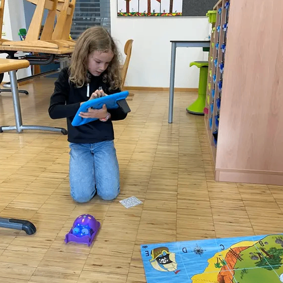 Ein Mädchen kniet auf dem Boden, hält ein Tablet und spielt ein Spiel. Ein Kartenspiel liegt vor ihr auf dem Boden.