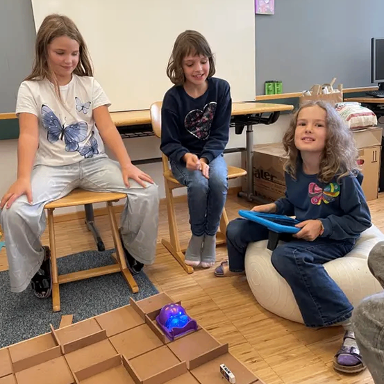 Drei Mädchen sitzen auf Stühlen und einem Beanbag-Sessel und lächeln in die Kamera. Ein labyrinthartiges Spielaufbau steht auf dem Boden, mit einem blauen Licht in der Mitte. Der Raum hat Holzböden, eine weiße Wand und Tische mit verschiedenen Gegenständen.