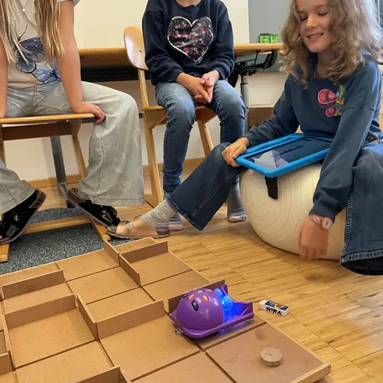 Drei junge Mädchen sitzen auf Stühlen und eine auf einem Balanceball und spielen mit einem kleinen Roboter auf einem Labyrinthbrett auf einem Holzboden.