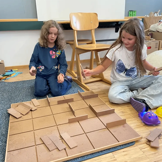 Zwei junge Mädchen sitzen auf dem Boden und beschäftigen sich mit einer Aktivität mit Kartonteilen. Eine hält ein kleines Stück, während die andere ein größeres benutzt. Sie sind von verschiedenen Kartonmaterialien und anderen Spielzeugen umgeben.