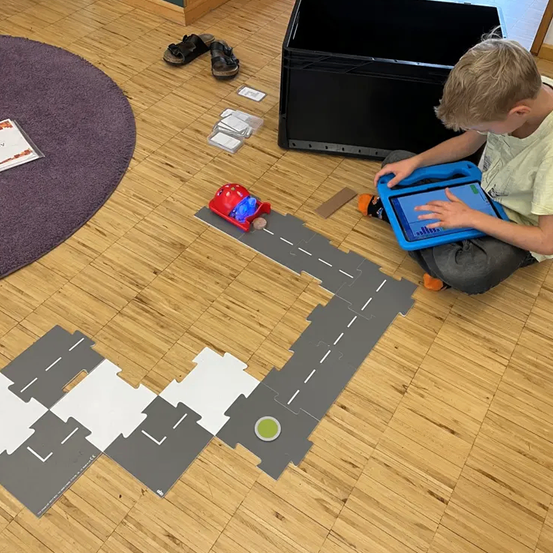 Ein junger Junge sitzt auf dem Boden und ist in ein Tablet vertieft, vor ihm liegt ein Puzzle-Weg. Ein rotes Spielzeugauto ist auf dem Puzzle platziert.