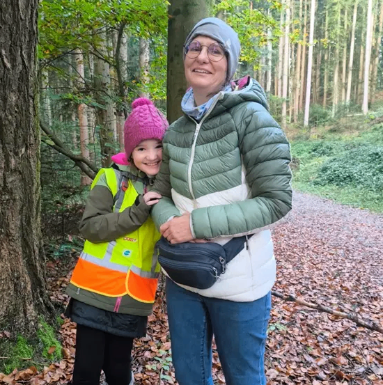 Eine erwachsene Frau und ein junges Mädchen stehen lächelnd zusammen im Wald. Das Mädchen trägt eine Warnweste, und die Frau trägt eine grüne Daunenjacke und eine graue Mütze.