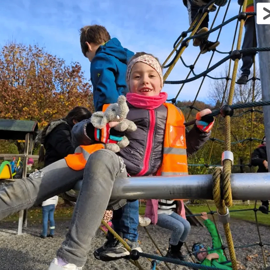 Kinder spielen auf einem Spielplatz, ein Mädchen hält ein Kuscheltier und klettert an einer Seilleiter.